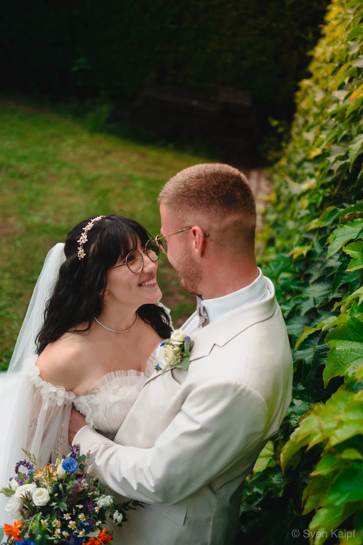 Hochzeitspaar-Portrait: Braut blickt lächelnd zum Bräutigam auf, Aufnahme von oben neben einer Efeuwand mit Blumenstrauß