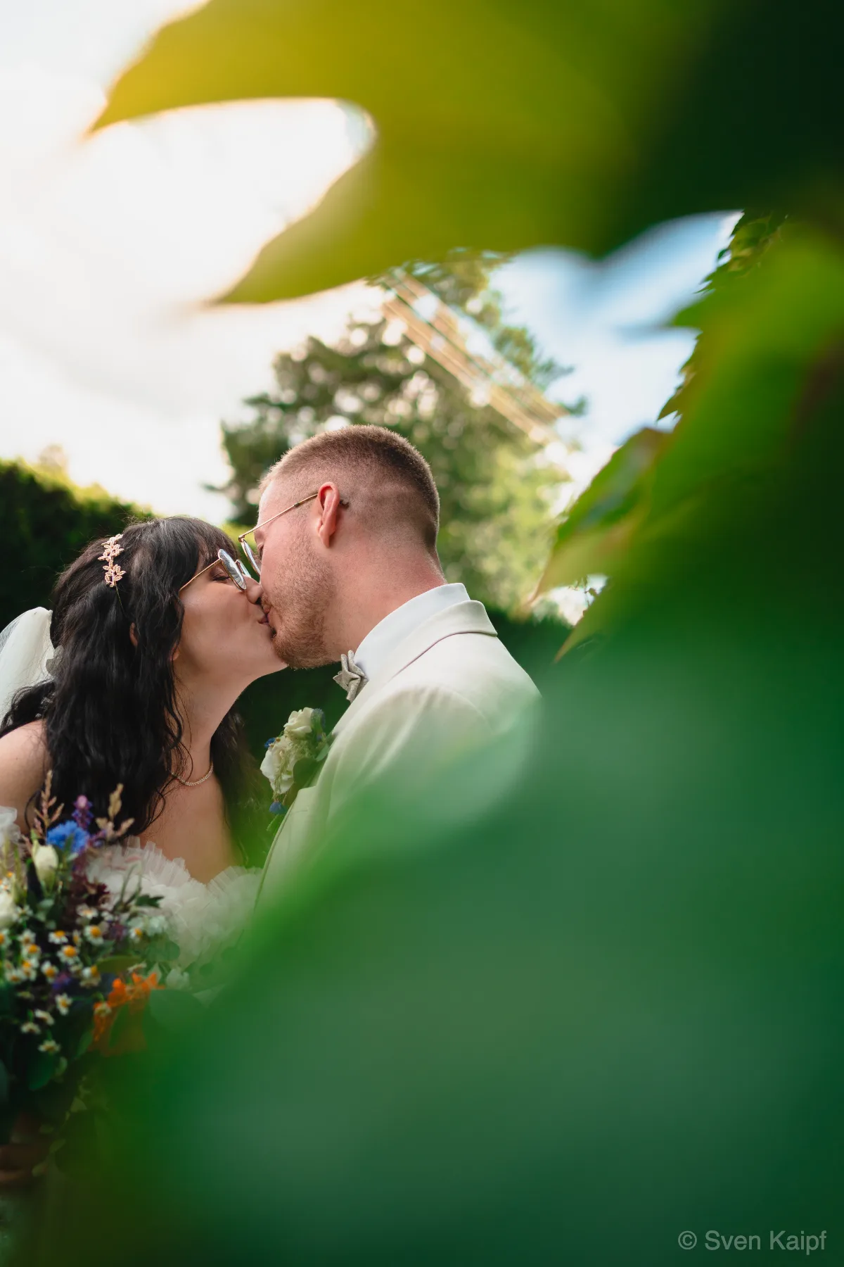 Hochzeitsfoto mit Kuss: Hochzeitspaar küsst sich, durch unscharfe grüne Blätter im Vordergrund fotografiert