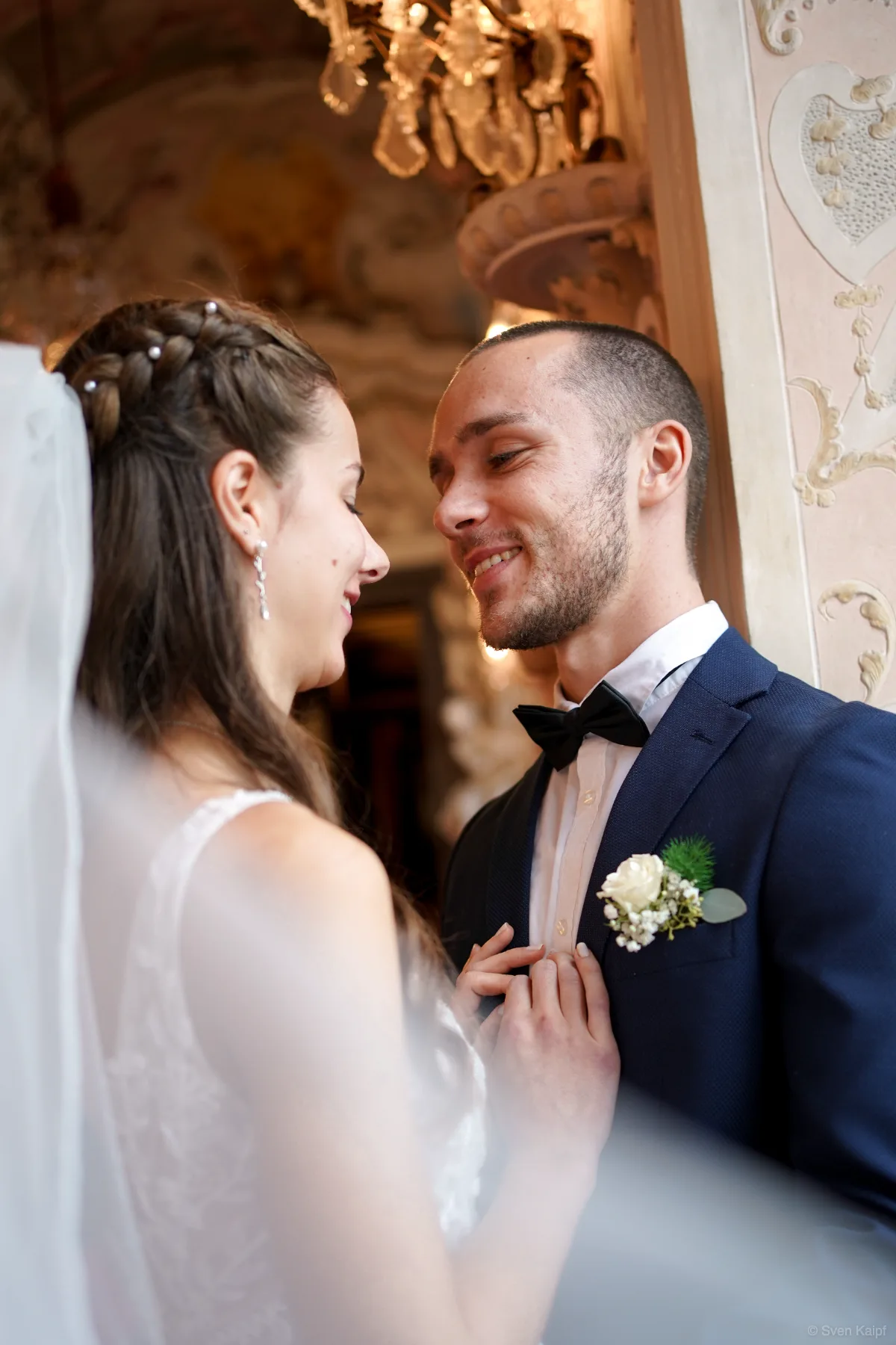 Hochzeitsportrait im barocken Saal: Hochzeitspaar lächelt sich an, Bräutigam mit Fliege und Blumenanstecker, Braut mit Schleier
