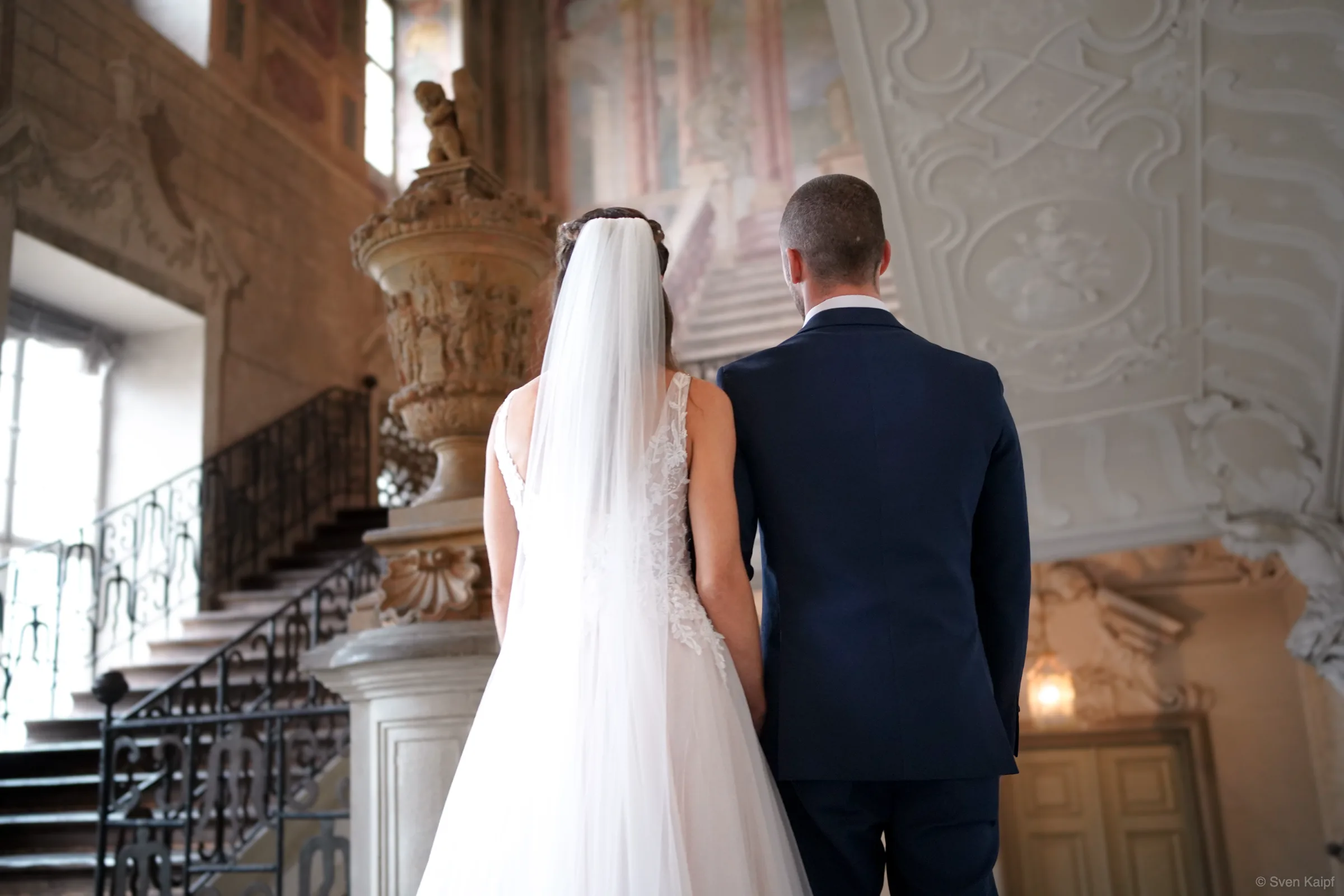 Hochzeitsbild von hinten: Hochzeitspaar im barocken Treppenhaus mit Stuckdecke, Fresken und schmiedeeisernem Geländer