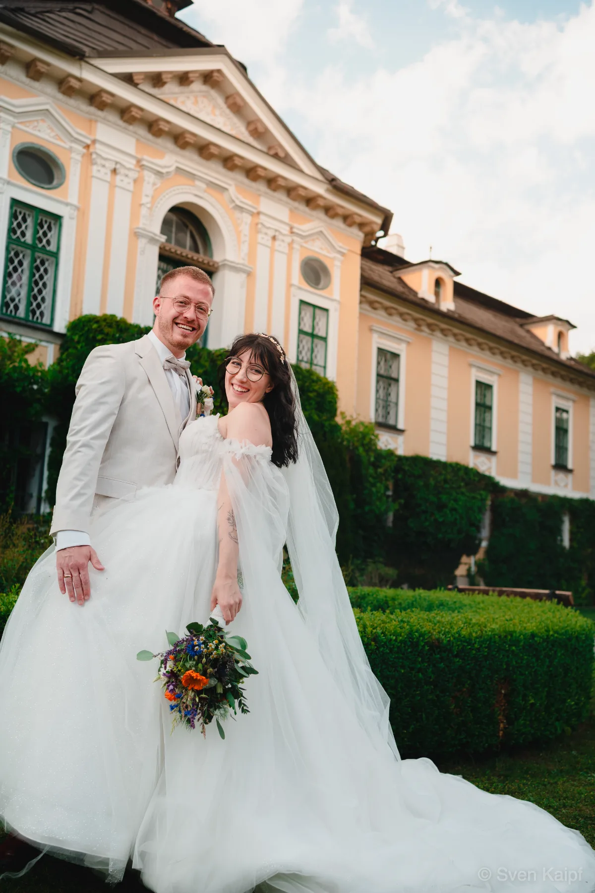 Hochzeitspaar vor einem gelben Barockschloss, Hochzeitsfoto mit Braut im langen Schleier und Blumenstrauß