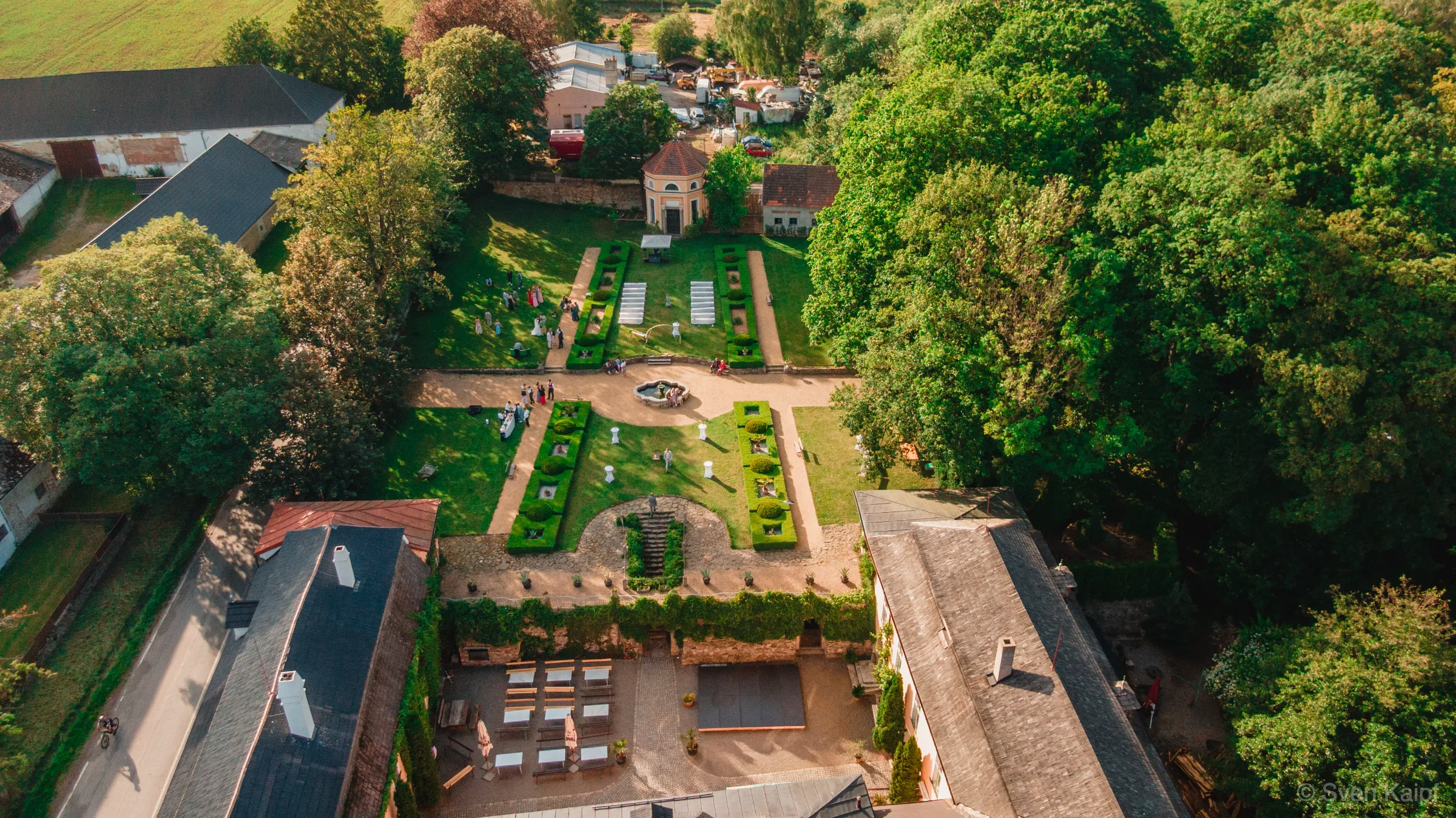 Drohnenaufnahme einer Schlosshochzeit: Schlossgarten mit Heckenbeeten, Springbrunnen und Hochzeitsgästen im Abendlicht